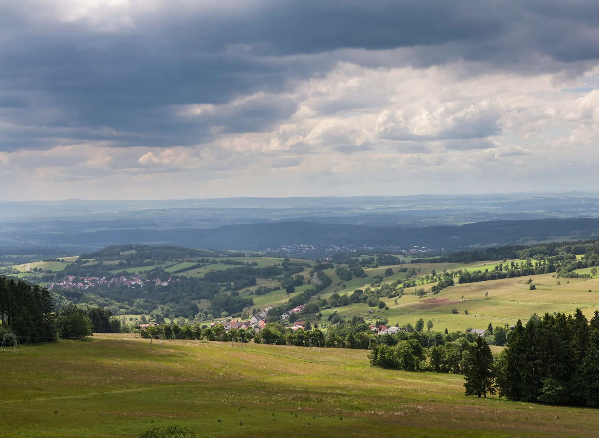 Fotografie einer weitläufigen Hügellandschaft mit grünen Wiesen, verstreuten Bäumen und kleinen Dörfern im Tal unter einem bewölkten Himmel. Die Szene zeigt Kontraste zwischen sonnendurchfluteten Flächen und schattigen Bereichen, was auf wechselhaftes Wet