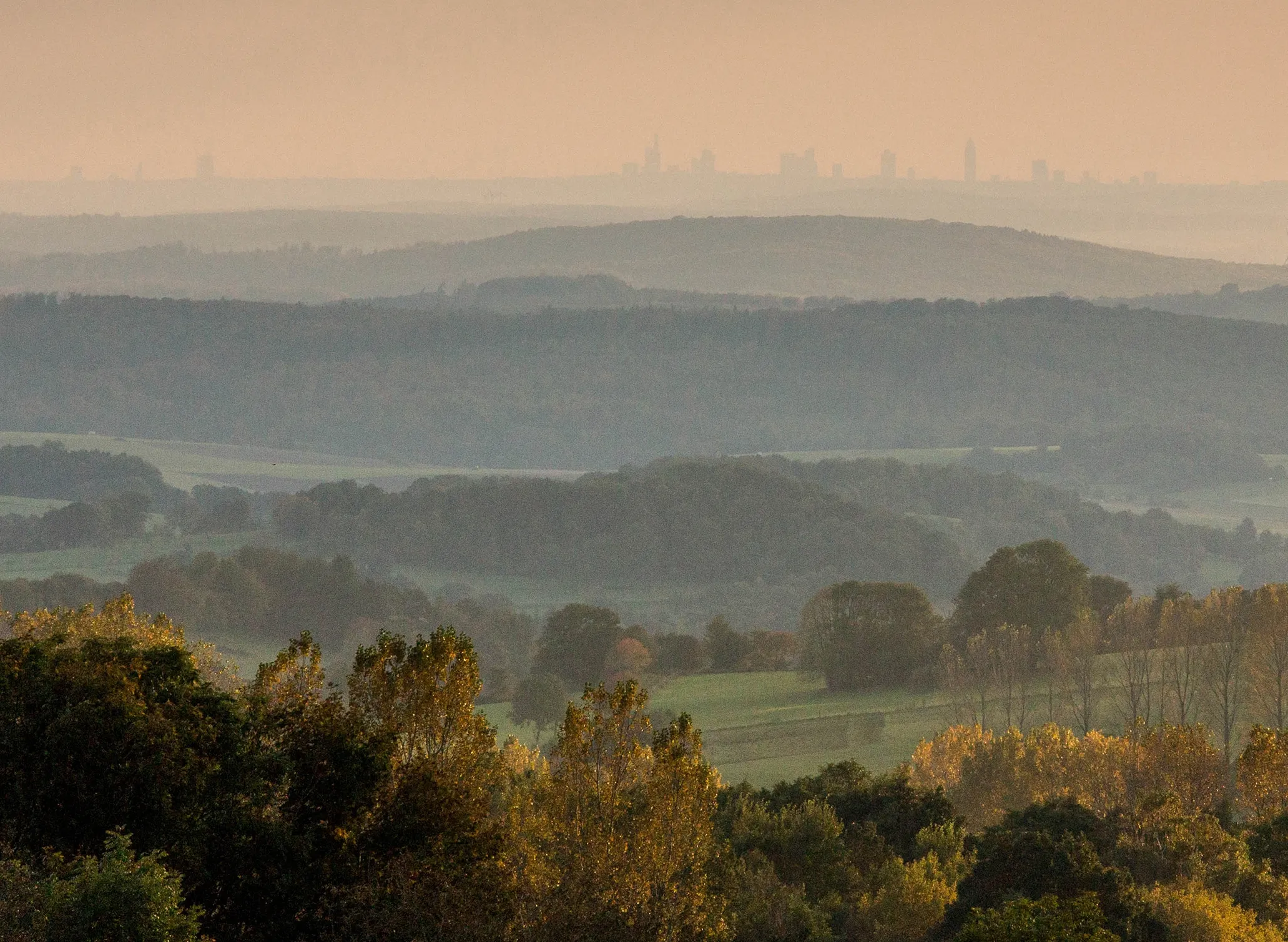 Fotografie einer herbstlichen Landschaft mit mehrschichtigen Wald- und Feldflächen im Vordergrund und einer nebligen Stadtsilhouette am Horizont. Sanfte Farbverläufe von Grün zu Braun und ein orangefarbener Himmel betonen Tageszeit und atmosphärische Tief
