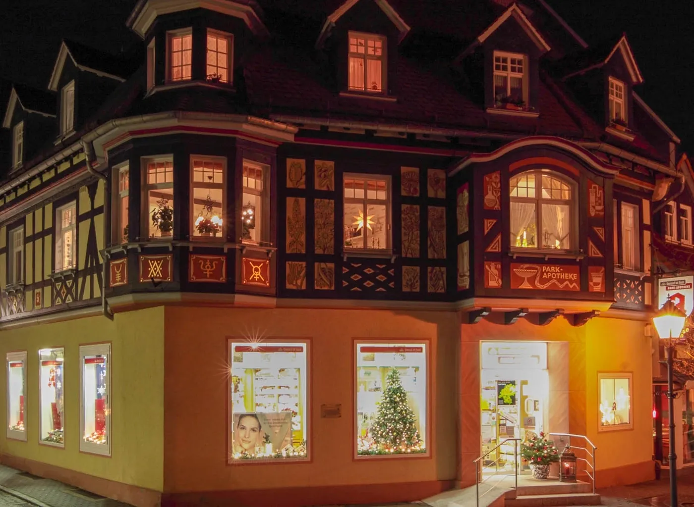 Fotografie eines historischen Fachwerkhauses bei Nacht mit beleuchteten Fenstern und weihnachtlicher Dekoration, einschließlich eines geschmückten Weihnachtsbaums im Schaufenster. Gebäude zeigt warmes Licht, sichtbare Details wie Holzverzierungen und die 
