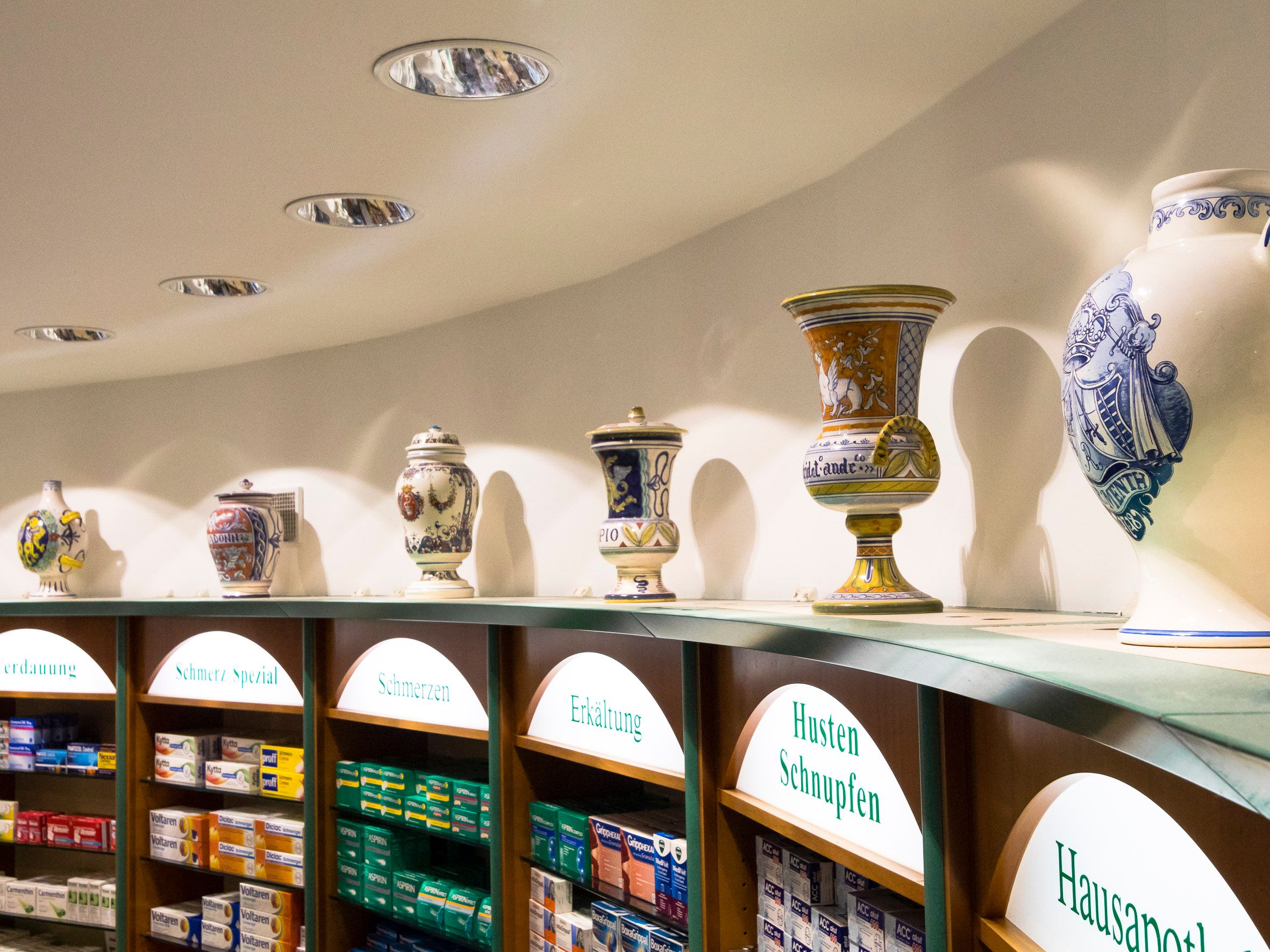 Fotografie einer Apothekeneinrichtung mit historischen Apothekengefäßen auf einem hohen Regal und darunter modernen Medikamenten in Regalfächern. Beschriftungen über den Fächern zeigen Kategorien wie "Erkältung" und "Husten Schnupfen", was auf eine themat
