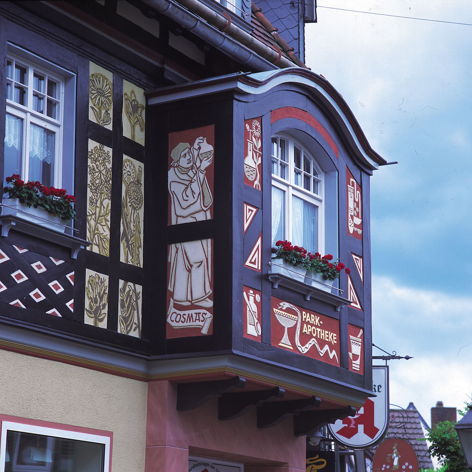 Fassadendetail eines Fachwerkhauses mit bemalten Holztafeln und dekorativen Fenstern, darunter ein Erker mit roten Blumen in Blumenkästen. Bemalungen zeigen historische Figuren, Pflanzenmotive und Symbole einer Apotheke, darunter Schriftzug „Park-Apotheke