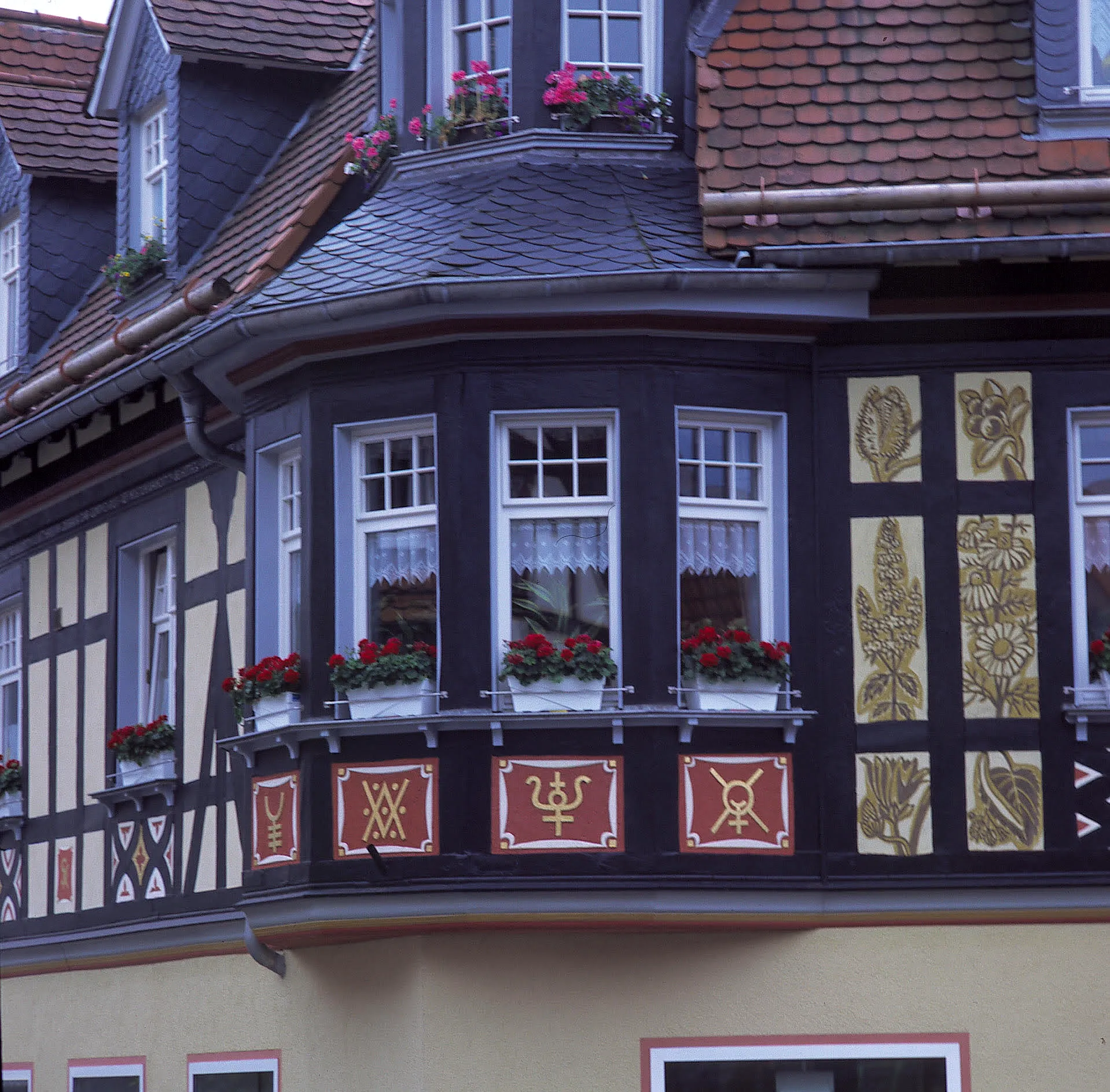 Fachwerkhaus mit Erker und dekorativen Holztafeln in Rot und Gold, die verschiedene Symbole und florale Motive zeigen. Fensterbänke sind mit roten Blumen geschmückt.